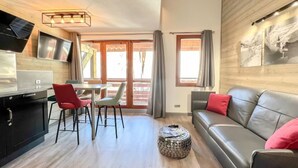 Una sala de estar moderna con un sofá, una mesa de comedor con sillas, una zona de cocina con un televisor y un ventanal con cortinas.
