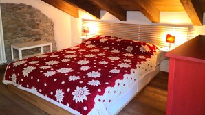 Une chambre avec un plafond en bois, un mur en pierre, un lit recouvert d’une courtepointe à motifs floraux rouges, des tables de chevet avec des lampes et une petite table blanche.