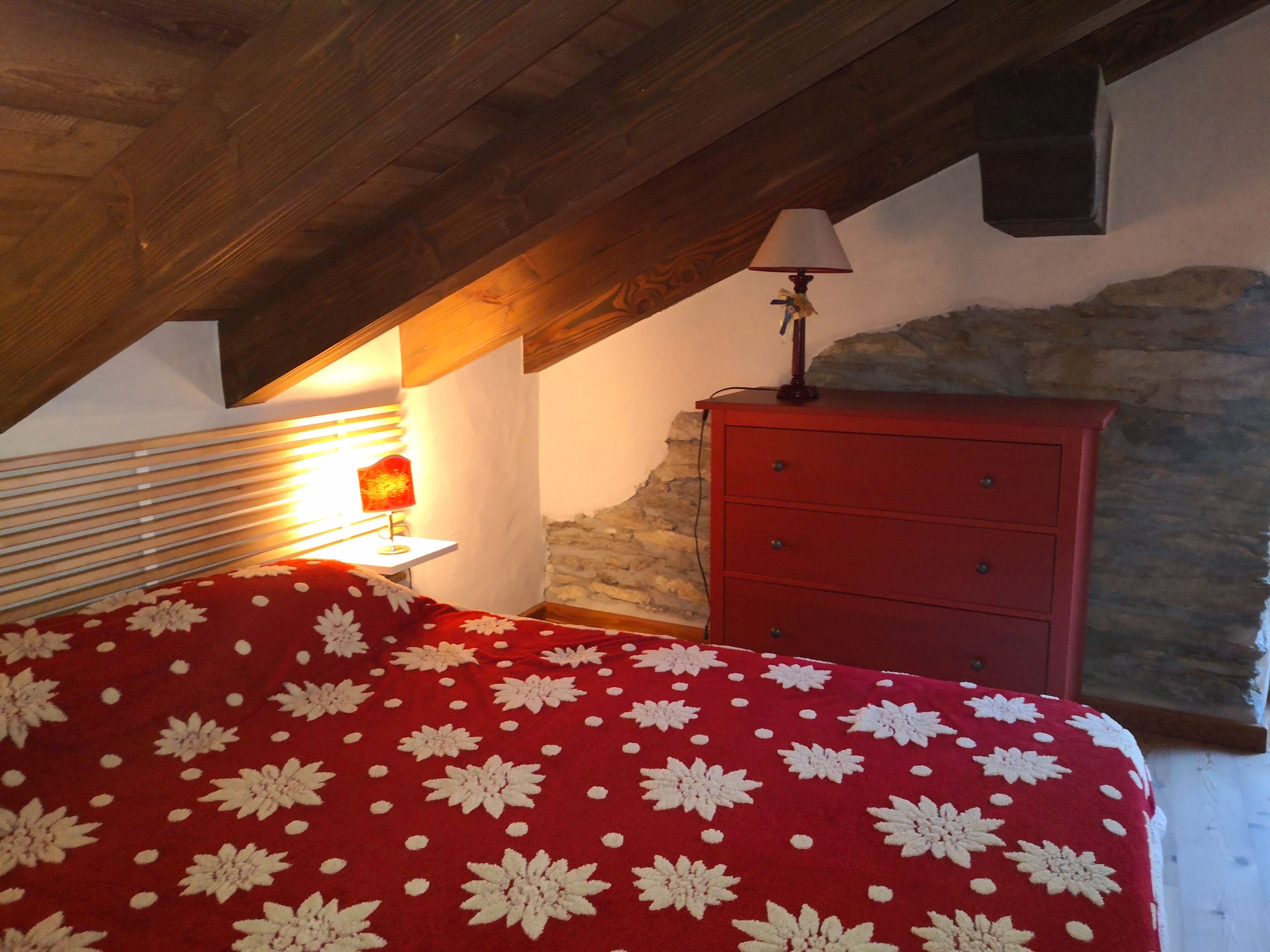 Een slaapkamer met een schuine plafond, een rode bed met wit bloemenbeddels, een nachtkastje met een lamp, en een rode kledingkast tegen een stenen muur.