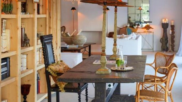 Uma sala de jantar com uma mesa de madeira, cadeiras e um candelabro. Há uma estante cheia de livros e objetos decorativos.