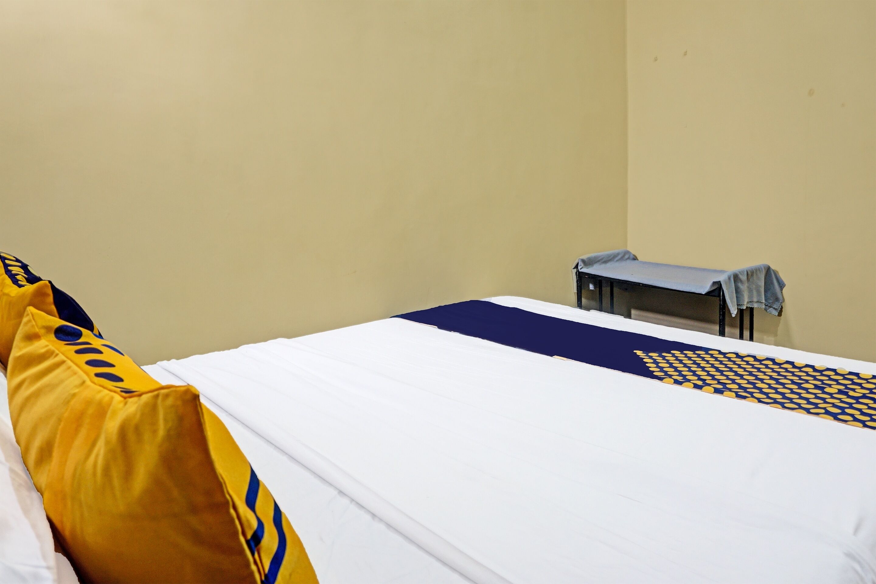 Una cama individual con una colcha blanca y azul, un almohadón amarillo y una mesita con mantel azul.