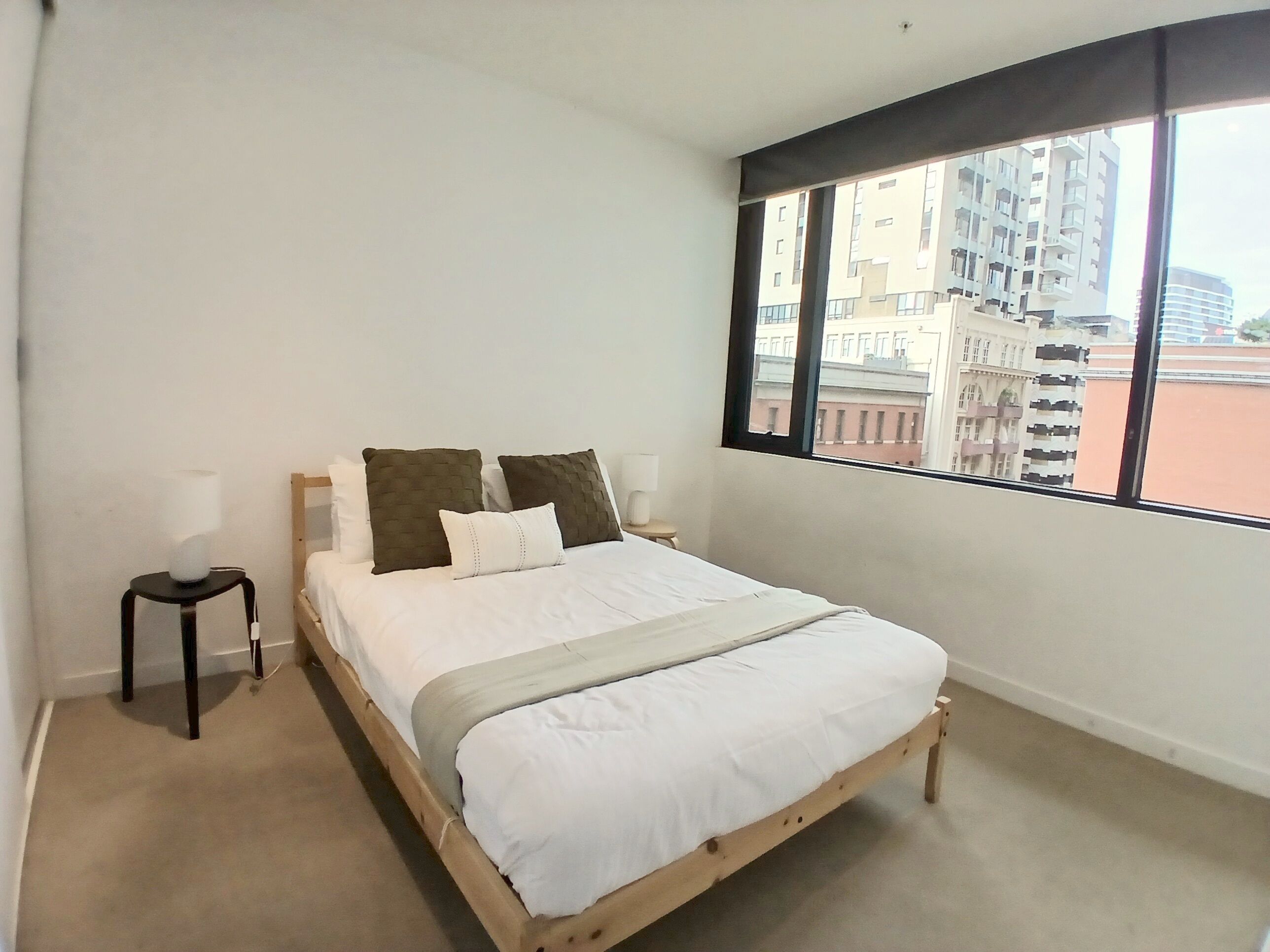 A bedroom with a bed, a nightstand, a lamp, and a large window with a view of buildings.
