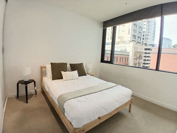 A bedroom with a bed, a nightstand, a lamp, and a large window with a view of buildings. - ReadySet Apartment at Midtown (Melbourne)