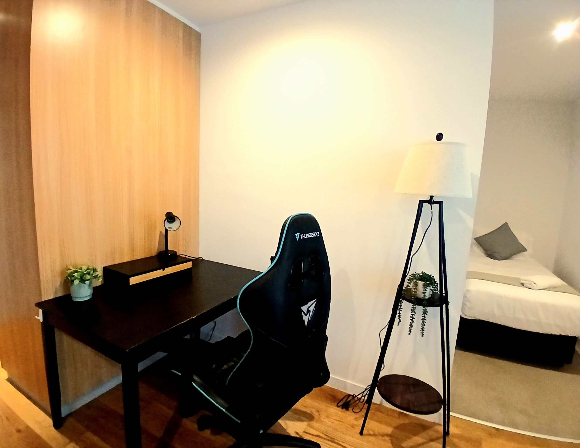 A modern room with a wooden wall, a desk with a computer and a chair, a lamp, and a bed with a pillow.