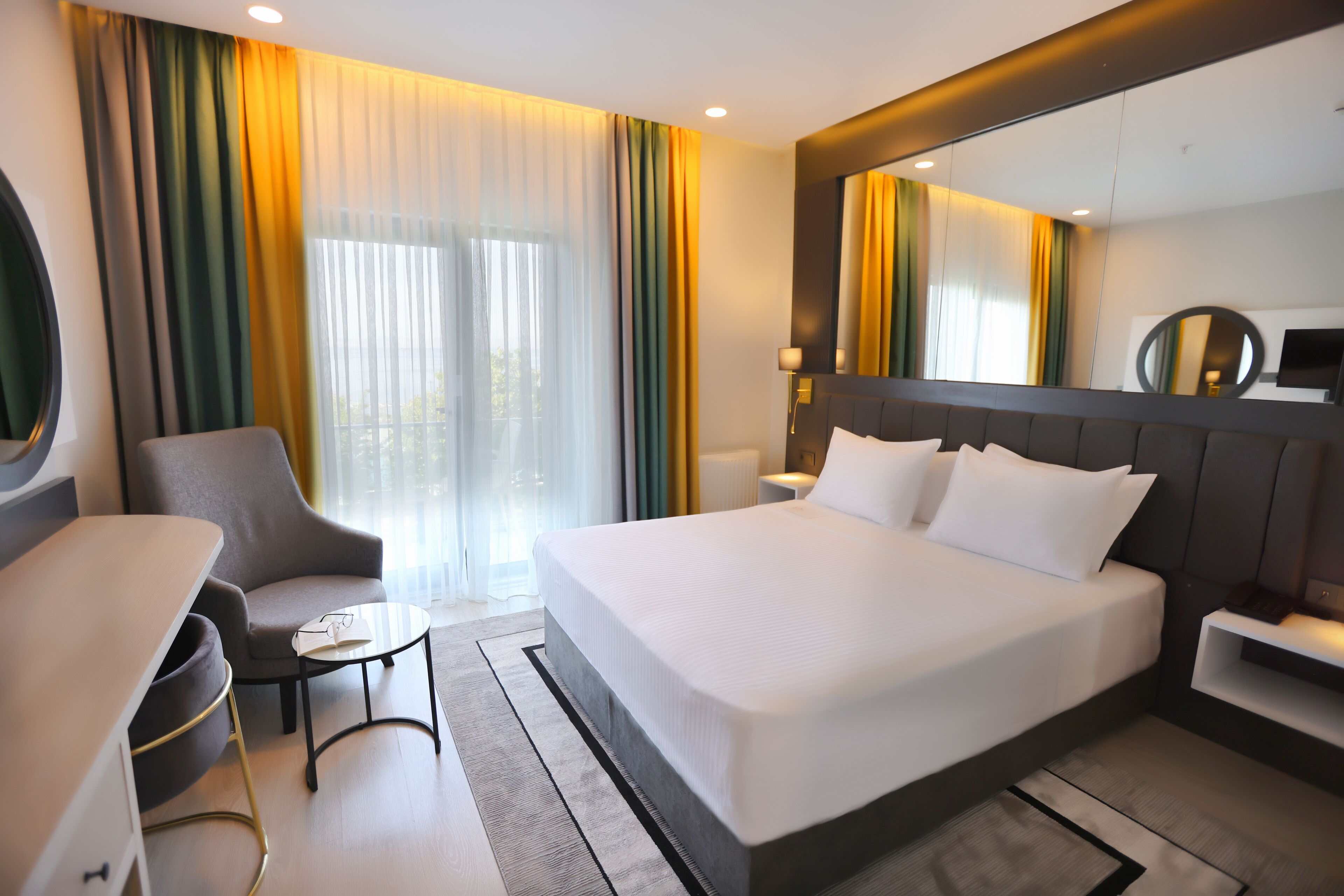 A modern hotel room with a large bed, a desk, a chair, a small table, and a large window with curtains.