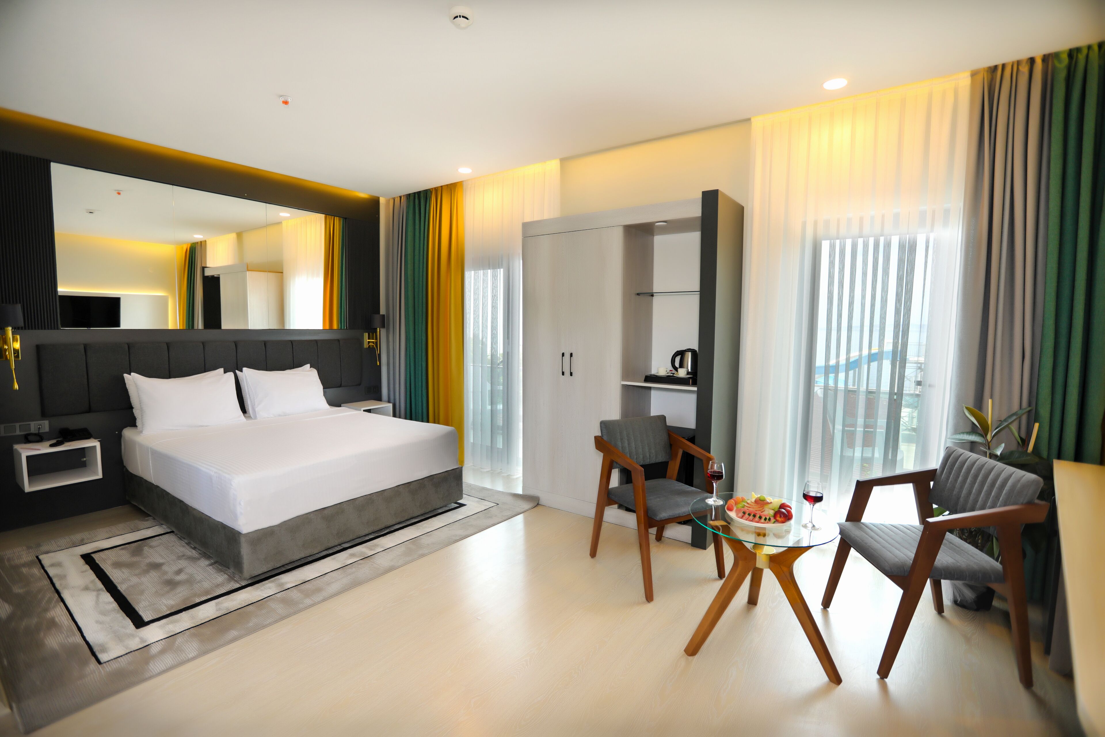 A modern hotel room with a large bed, a desk with a chair, a dining table with chairs, and a wardrobe.