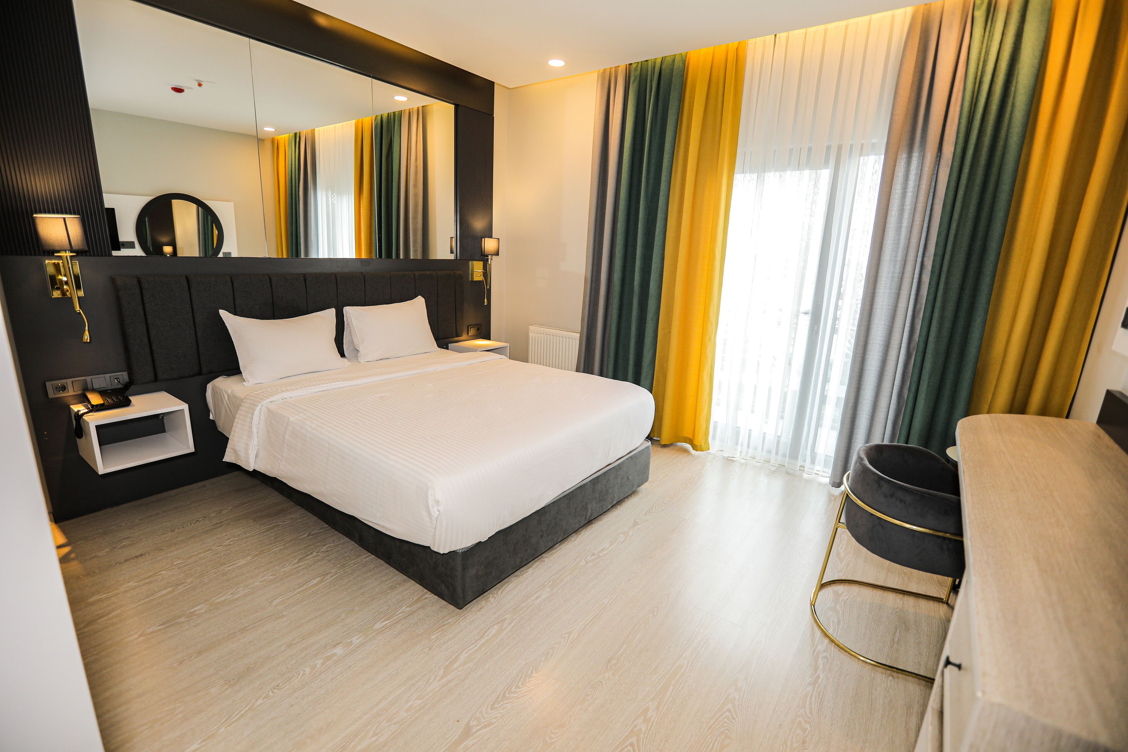 Una habitación de hotel moderna con una cama grande, mesitas de noche, un espejo y vistas al exterior a través de cortinas.