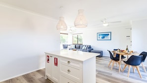 Una cocina moderna con armarios blancos, una isla central, luces colgantes y un comedor con mesa lista para cuatro.