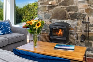 Ein gemütliches Wohnzimmer mit Kamin aus Stein, einer grauen Couch mit blauen Kissen und einem Strauß Sonnenblumen auf einem Holztisch.