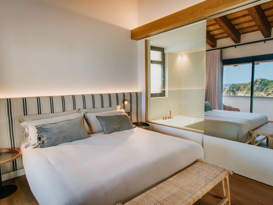 Ein Schlafzimmer mit einem großen Bett, einem Nachttisch, einem Spiegel und Blick auf einen Balkon mit Tisch und Stühlen.