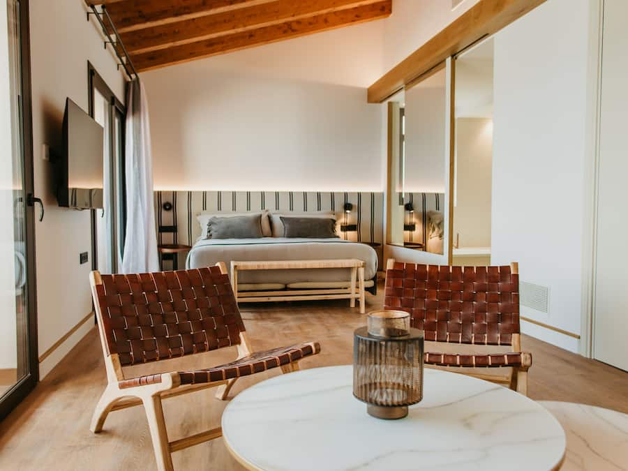 A modern hotel room with a wooden ceiling, a bed with a gray headboard, two leather chairs, a round marble table, and a television mounted on the wall.