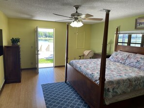 A four-poster bed with a floral bedspread, a ceiling fan, and a view of a pond through an open door. - Lake View Retreat Loaded For Fun, with STUNNING Views! (Inverness)