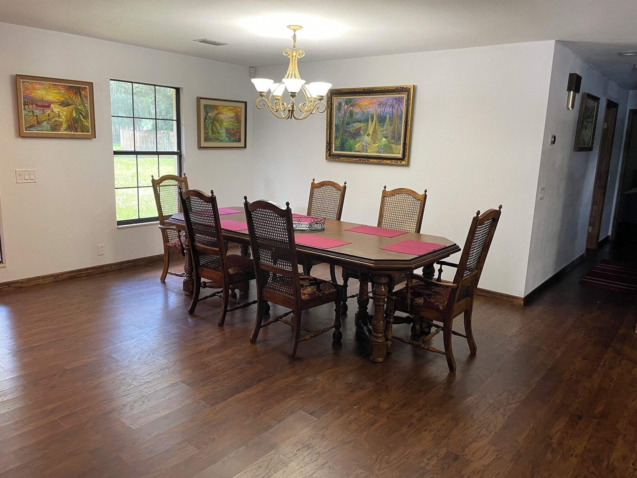 A dining room with a wooden table and chairs, a chandelier, and three paintings on the walls.