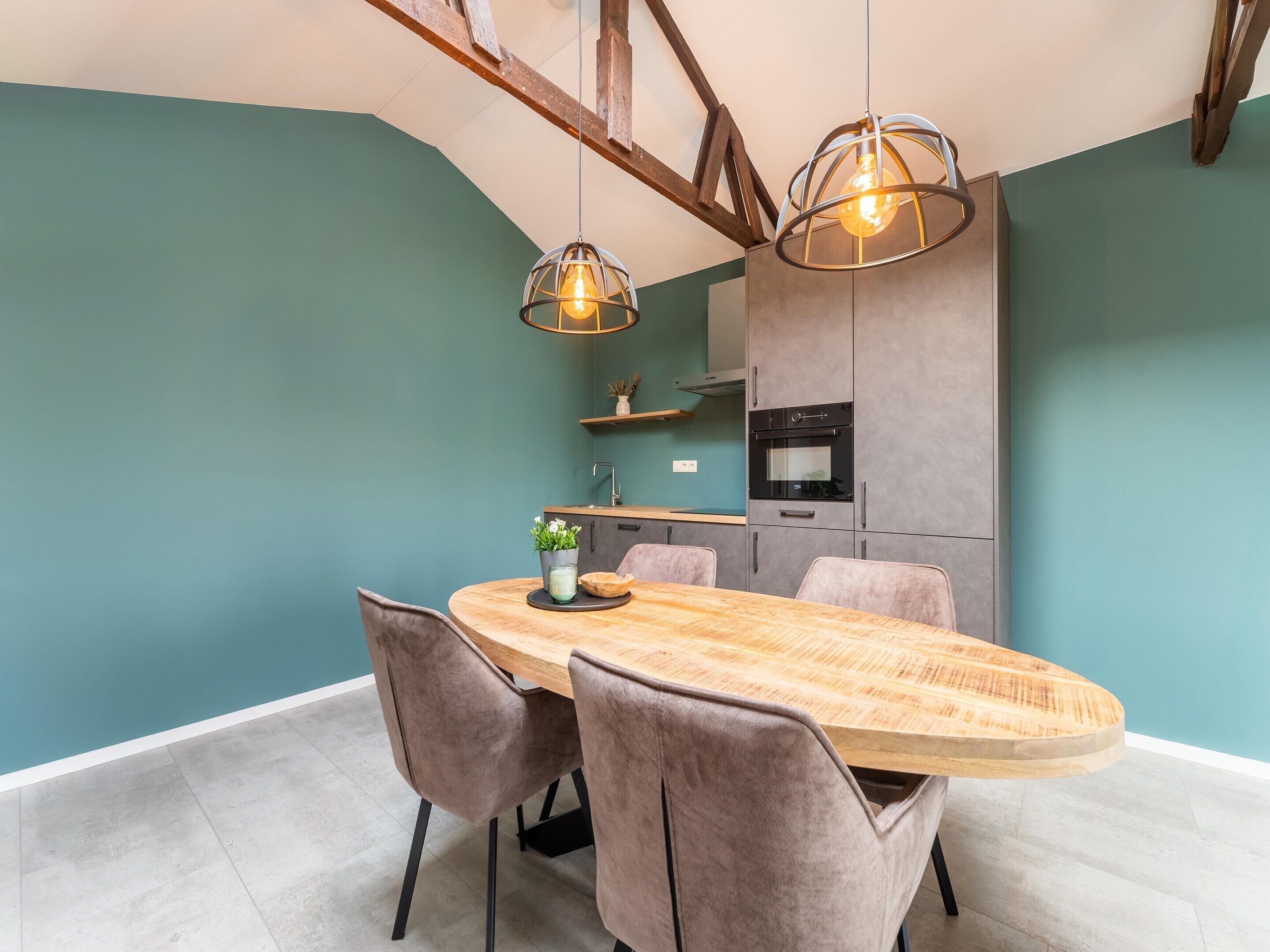 A dining area with a wooden table and cushioned chairs, a hanging light fixture, and a kitchen in the background.