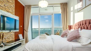 A modern bedroom with a large bed, a TV mounted on a stone wall, and a city view through large windows.
