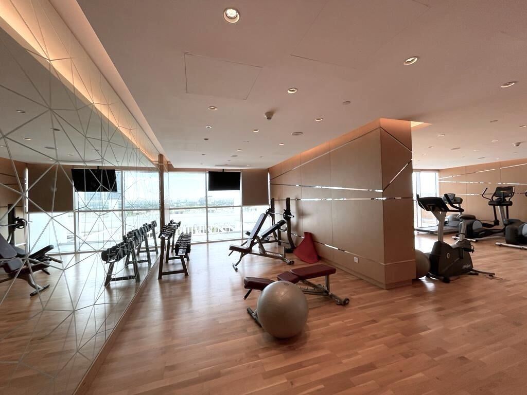 A modern gym with exercise equipment, large windows, and a wooden floor.