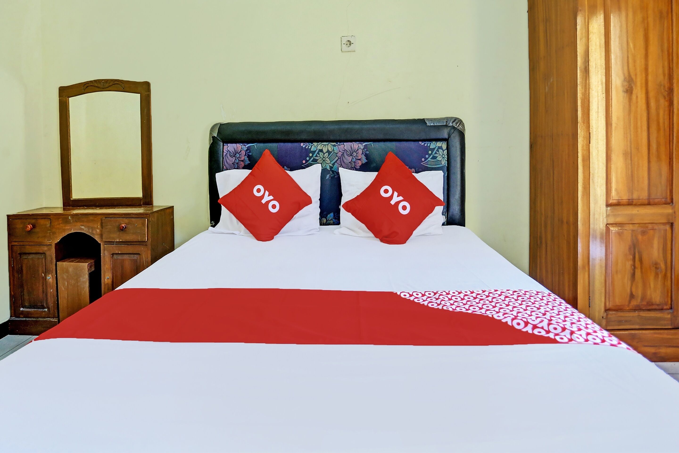 A bed with a red and white bedspread, two red pillows with "oyo" text, a wooden headboard, a wooden dresser with a mirror, and a wooden wardrobe.