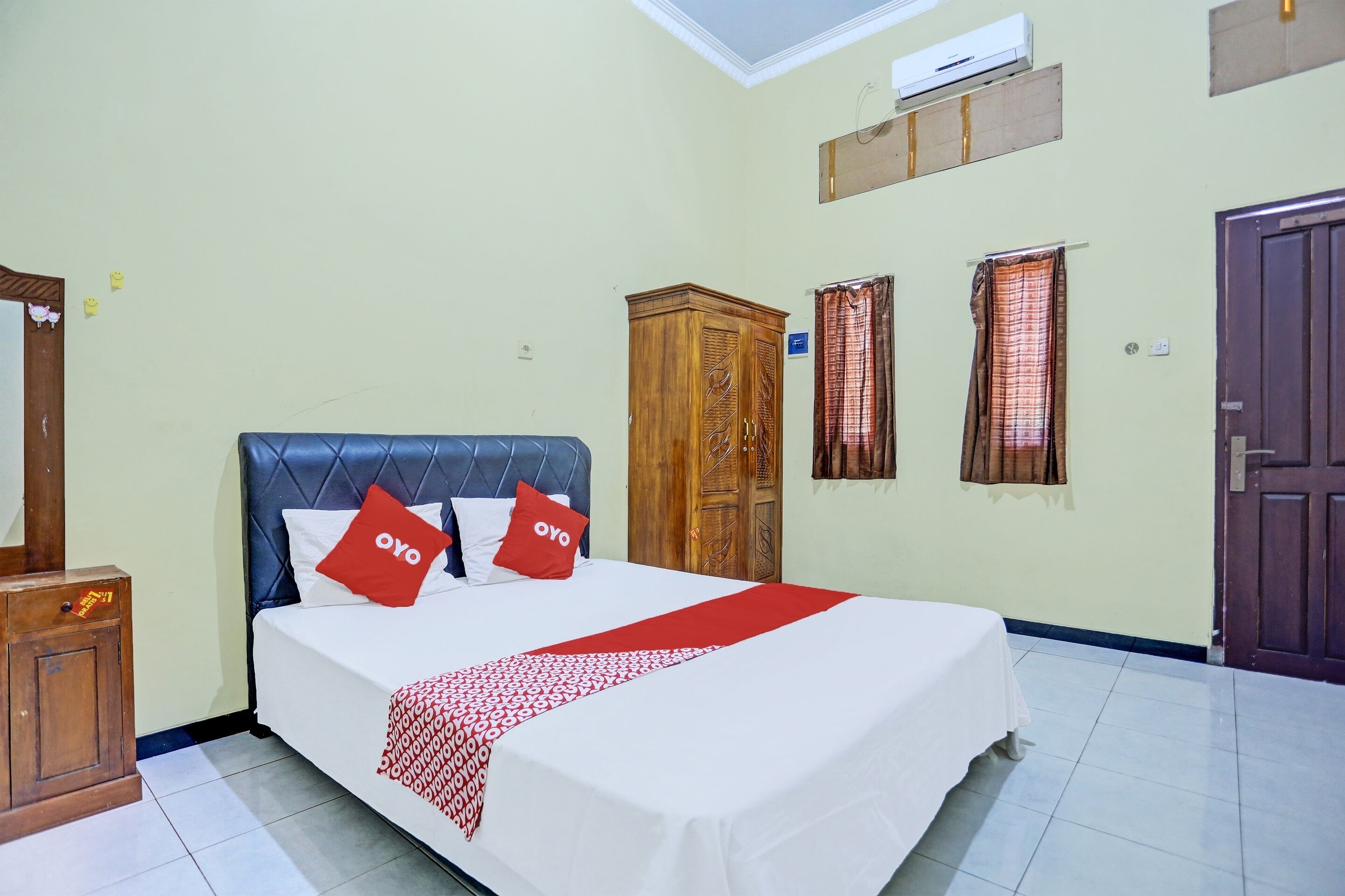 A hotel room with a bed, a wardrobe, a wardrobe mirror, a window with curtains, and an air conditioning unit.