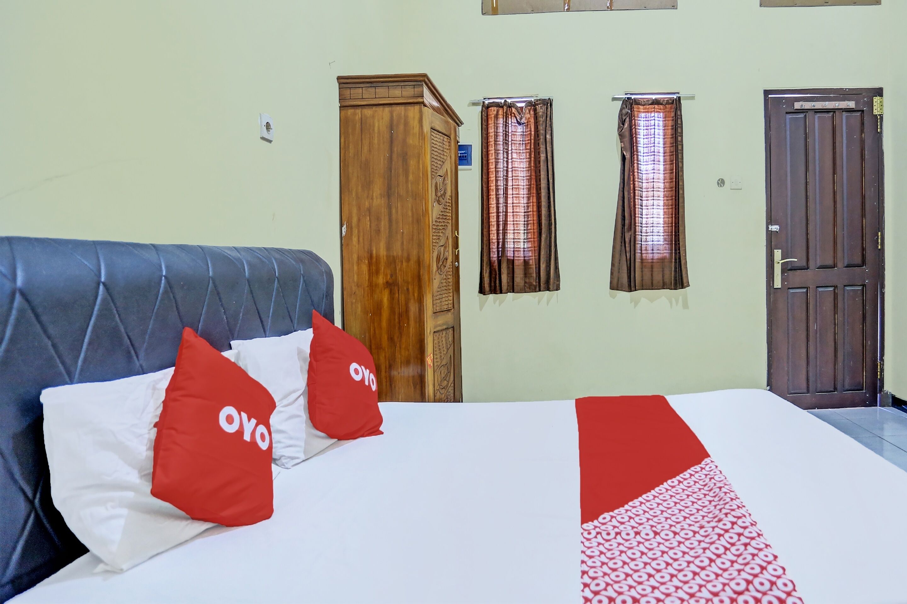 Habitación de hotel con una cama, dos almohadas rojas con el logotipo 'OYO', un armario de madera y dos ventanas con cortinas.