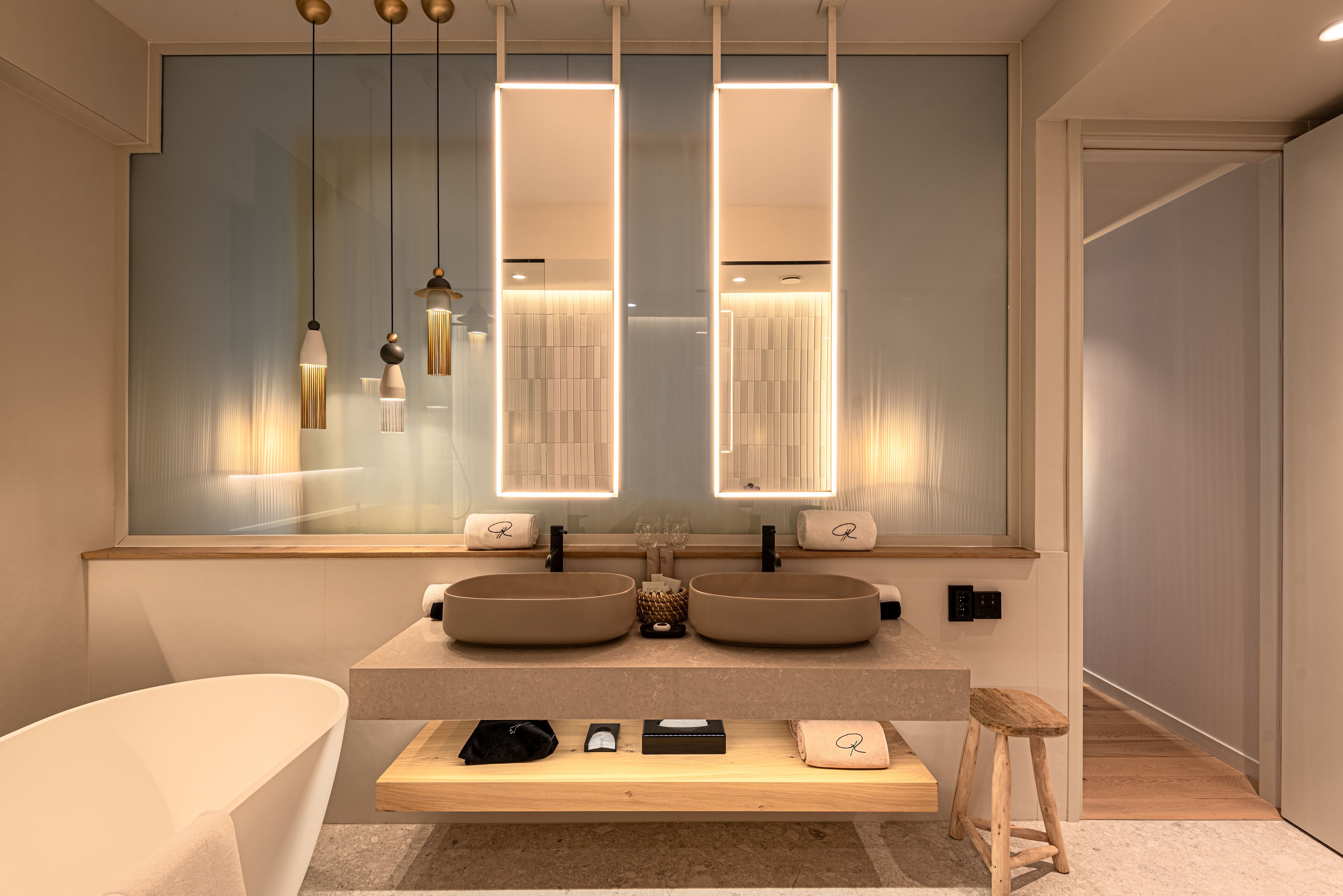 A modern bathroom with two sinks, a large mirror, pendant lights, and a wooden bench.