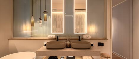 A modern bathroom with two sinks, a large mirror, pendant lights, and a wooden bench.