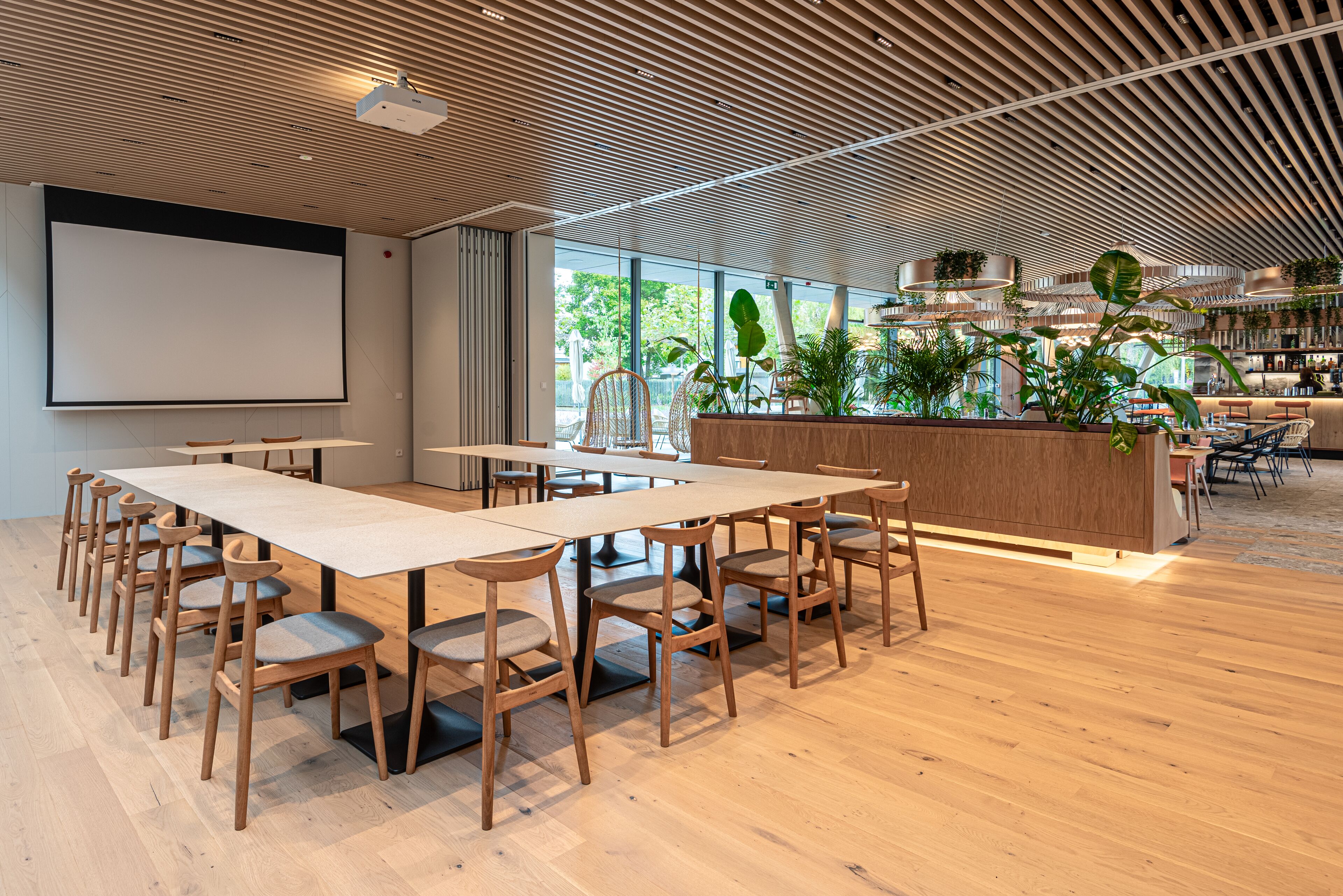 Een moderne vergaderruimte met een grote tafel, houten stoelen, een scherm voor een projecter en een receptiegedeelte met planten.
