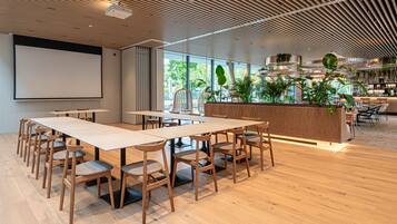 Une salle de conférence moderne, dotée d’une grande table, de chaises en bois, d’un écran de projection et d’un espace d’accueil agrémenté de plantes.