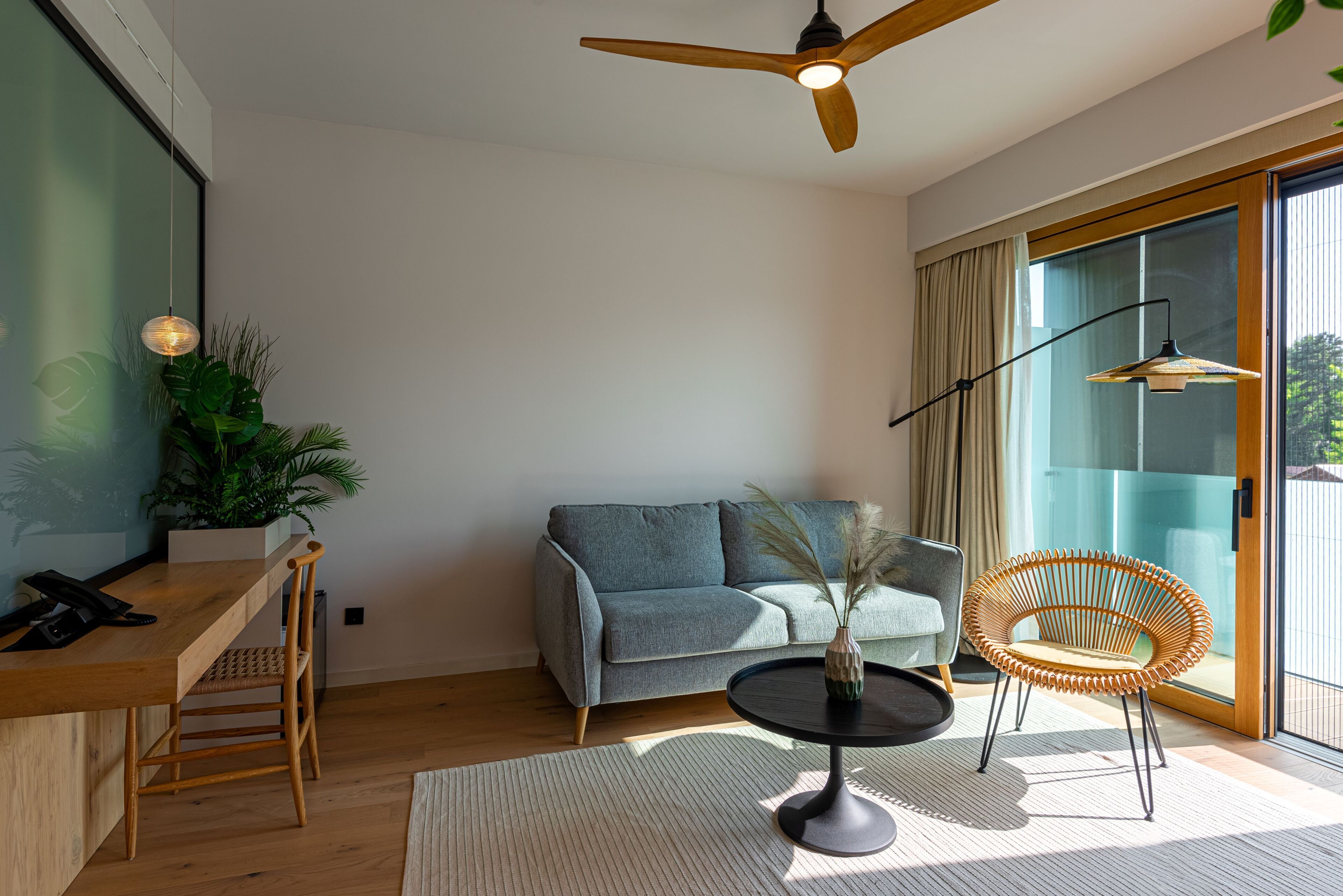 Un salon moderne avec un bureau en bois, un canapé gris, une table ronde ornée d’un vase, une chaise en osier et un ventilateur de plafond.