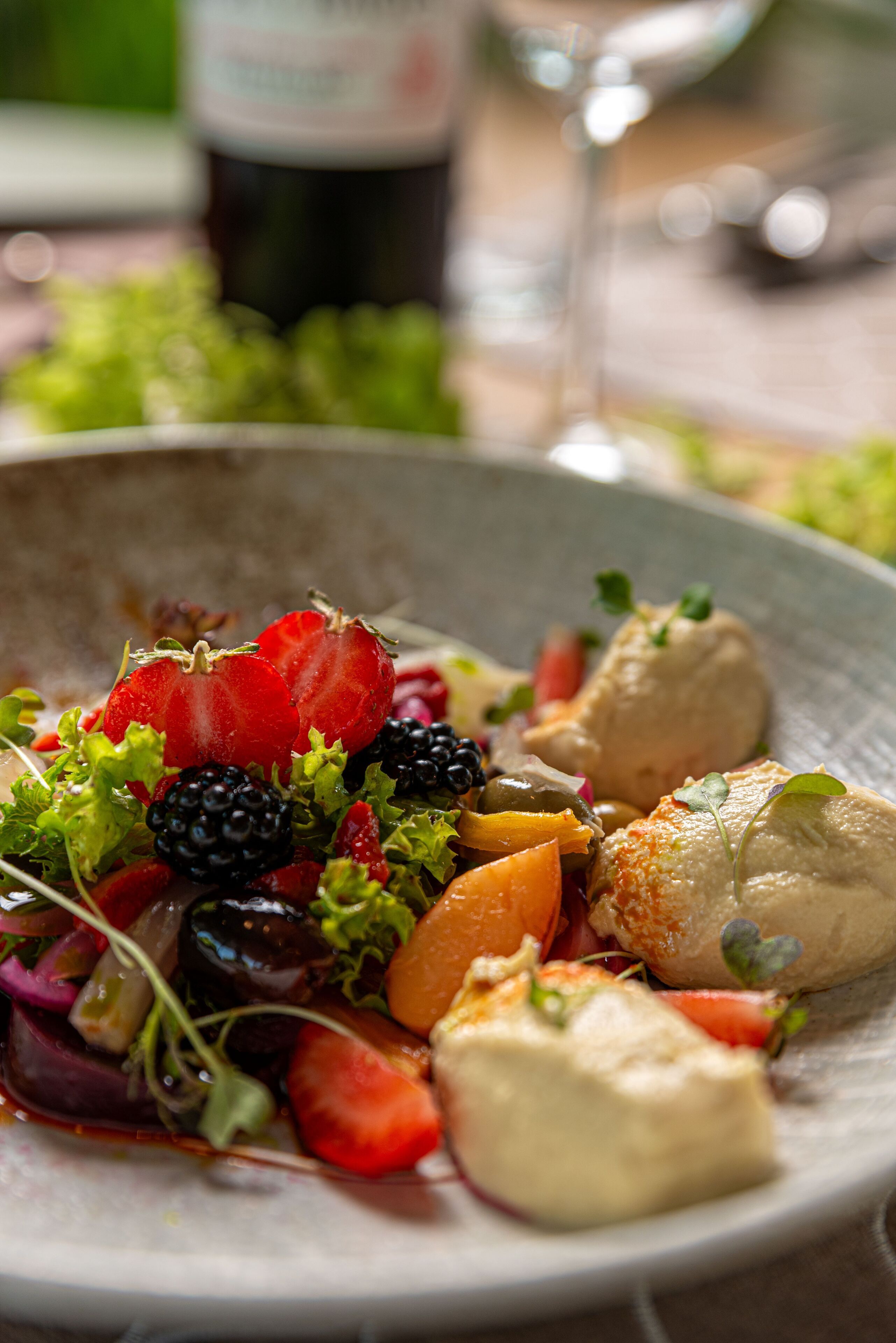 Um prato de comida com frutas e legumes variados, um copo de vinho e uma garrafa ao fundo.