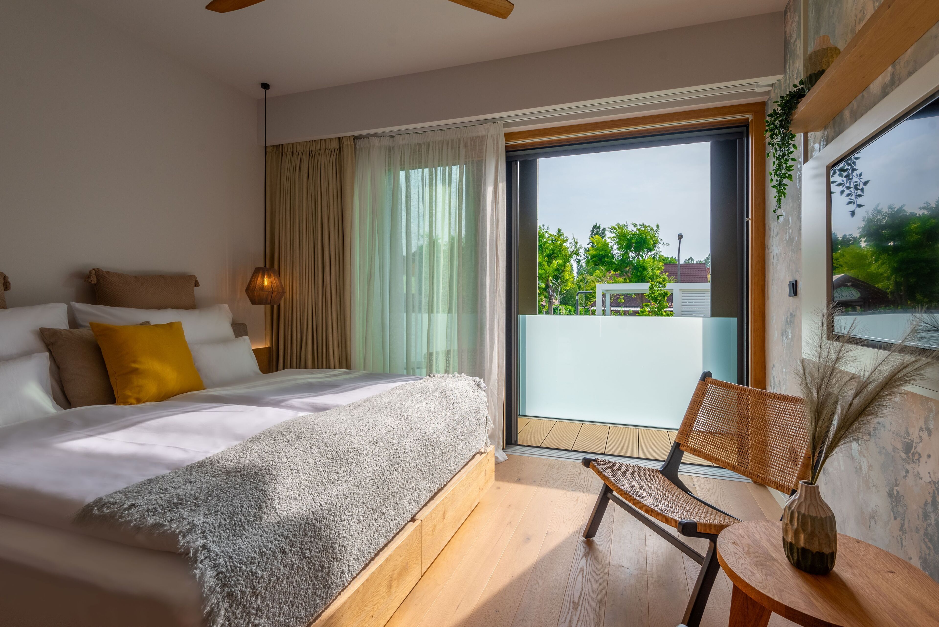 A modern bedroom with a large bed, a chair, and a balcony with a view of greenery.