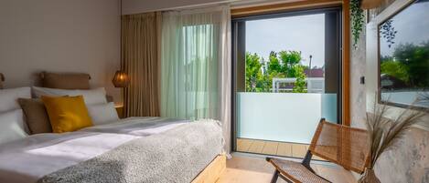 A modern bedroom with a large bed, a chair, and a balcony with a view of greenery.
