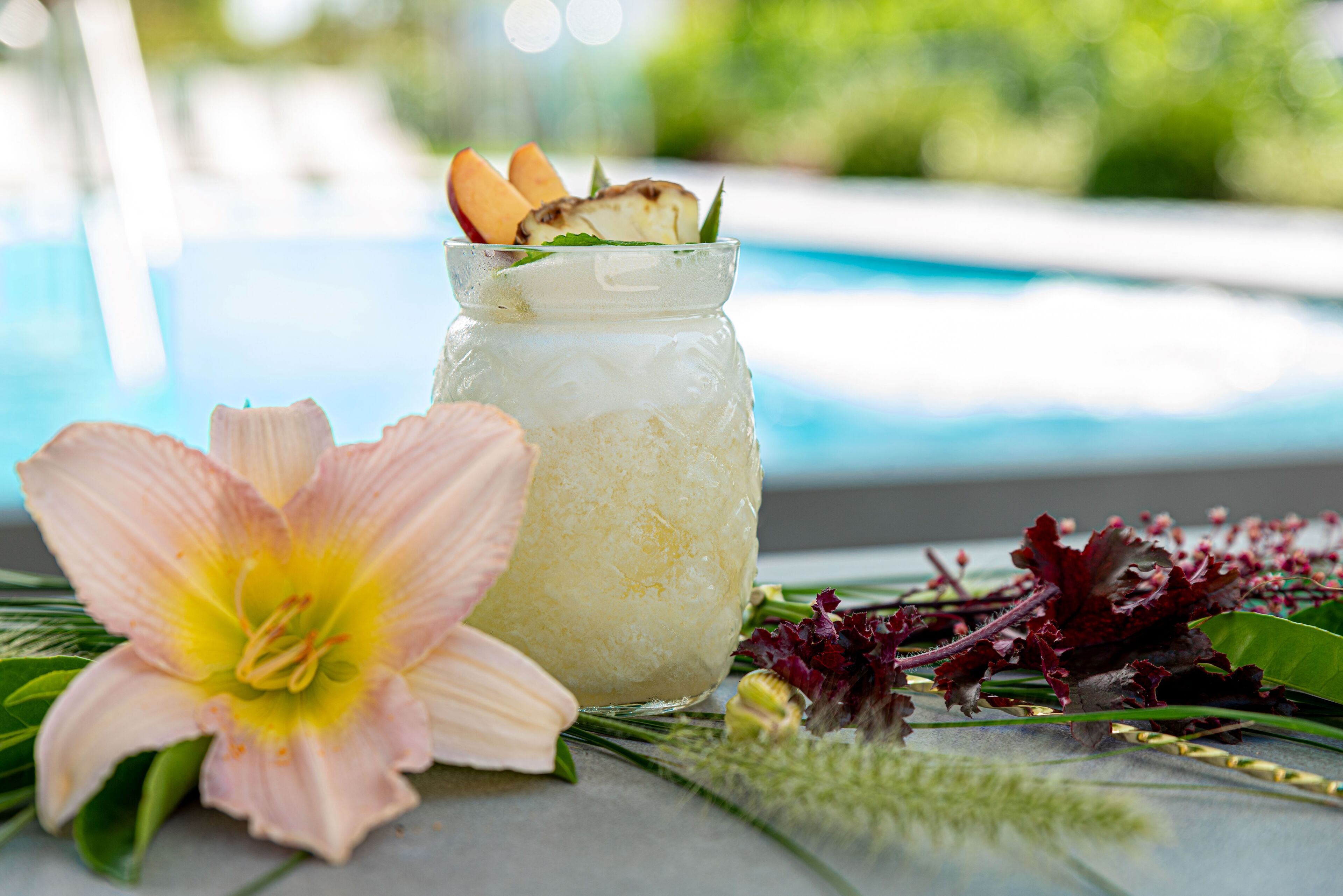 Un bocal en verre transparent contenant une boisson, une fleur rose et quelques feuilles vertes, placé à côté d’une piscine.