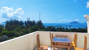 En balkon med bord og stole, der giver udsigt over kysten med en mole og et fjerntliggende bjerg.