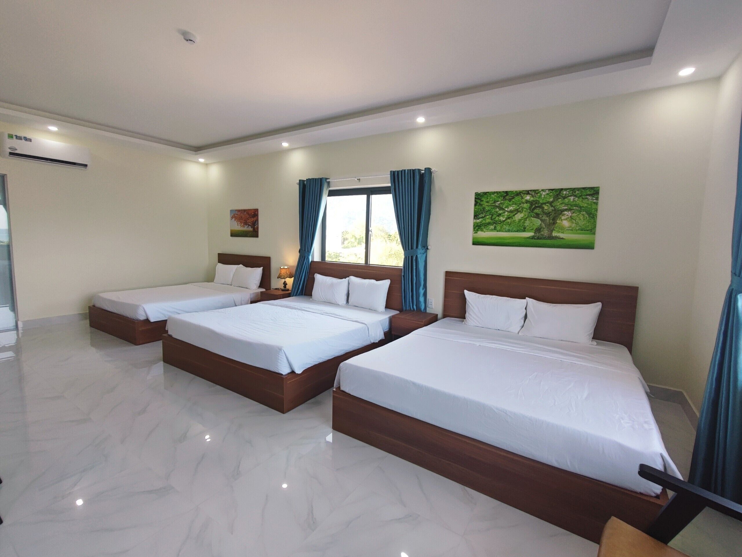 A hotel room with three beds, a large window with curtains, a painting on the wall, and a ceiling light.