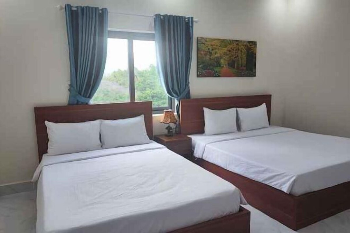 Une chambre d’hôtel avec deux lits, une fenêtre avec des rideaux, un tableau au mur et une table de chevet avec une lampe.