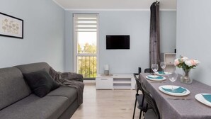 A modern room with a grey sofa, a dining table set for two, a flat-screen TV, and a window with blinds.