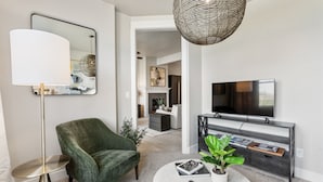 Un salon moderne avec un fauteuil vert, une table basse ronde sur laquelle sont posés des magazines et une plante, un lampadaire, un grand miroir, un téléviseur sur une console sombre et un luminaire suspendu en osier.