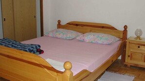 Een houten bed met een roze sprei en bloemenkussens, een nachtkastje van hout met een lamp, en een houten garderobe.