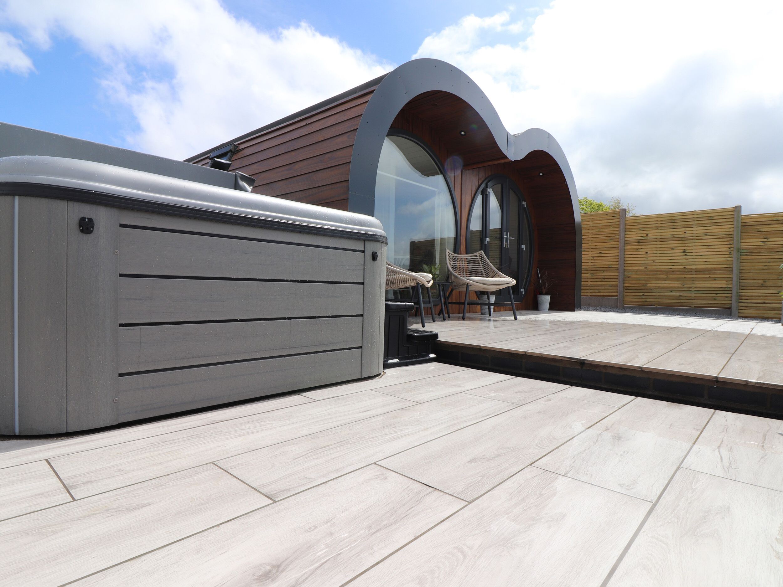 A modern outdoor patio with a hot tub, two wicker chairs, and a wooden fence.