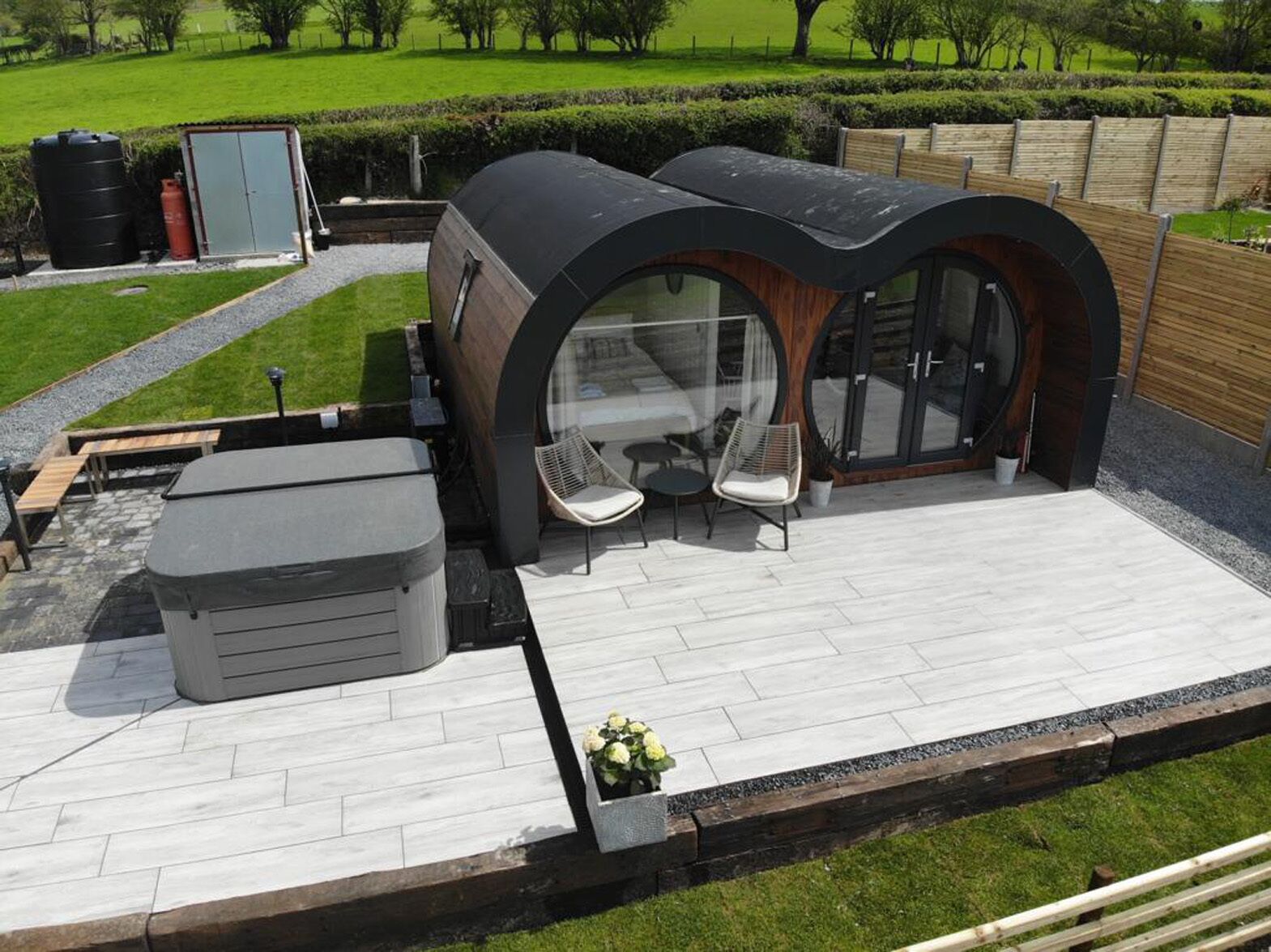 A modern outdoor patio with a curved glass enclosure, a hot tub, and a seating area with two chairs.