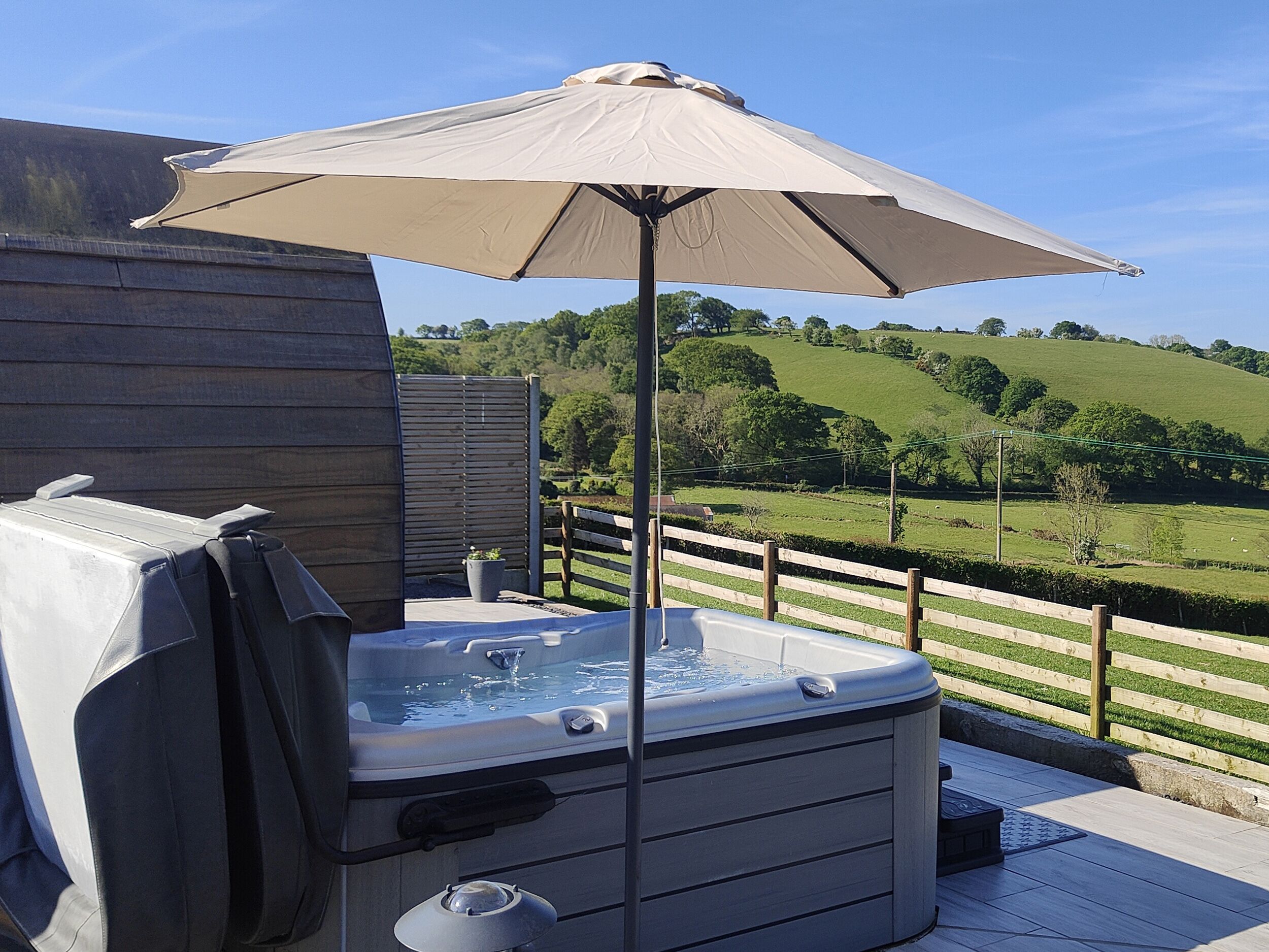 A hot tub with an umbrella on a wooden deck overlooking a green landscape.