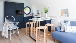 A compact kitchen with a round window, a dining area with stools, a white chair, and a blue sofa with patterned cushions. - Ceunant (Aberystwyth)