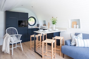 A compact kitchen with a round window, a dining area with stools, a white chair, and a blue sofa with patterned cushions.