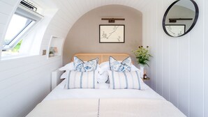 A bedroom with a curved ceiling, a bed with patterned pillows, a bedside table with a vase of flowers, a framed artwork on the wall, and a round mirror. - Ceunant (Aberystwyth)