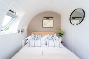 Un dormitorio con techo curvo, cama con almohadas estampadas, mesita de noche con un jarrón de flores, una obra de arte enmarcada en la pared y un espejo redondo.