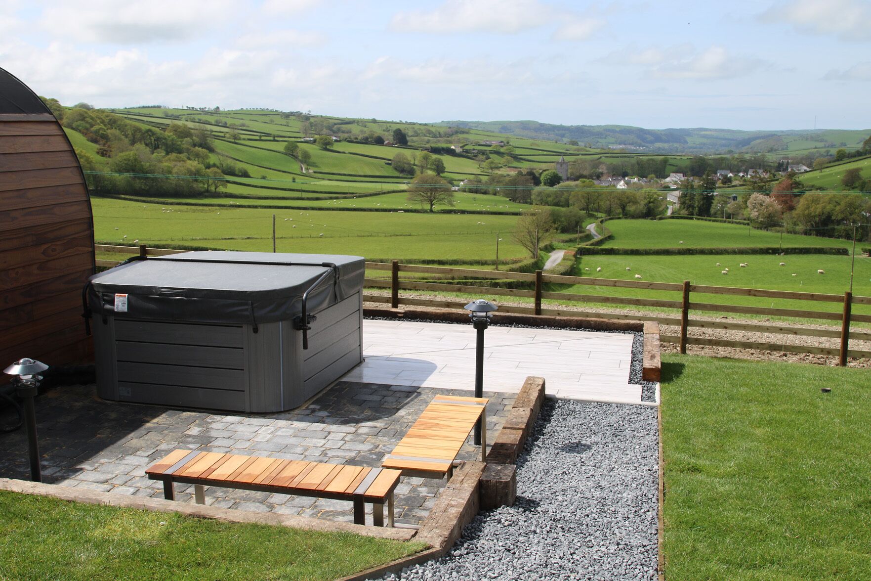 A patio area with a hot tub, wooden benches, and a view of rolling hills and a distant village.
