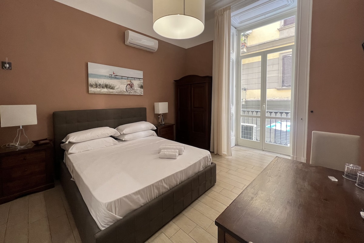 A hotel room with a bed, bedside tables, a lamp, a painting, an air conditioning unit, and a view of the outside balcony.