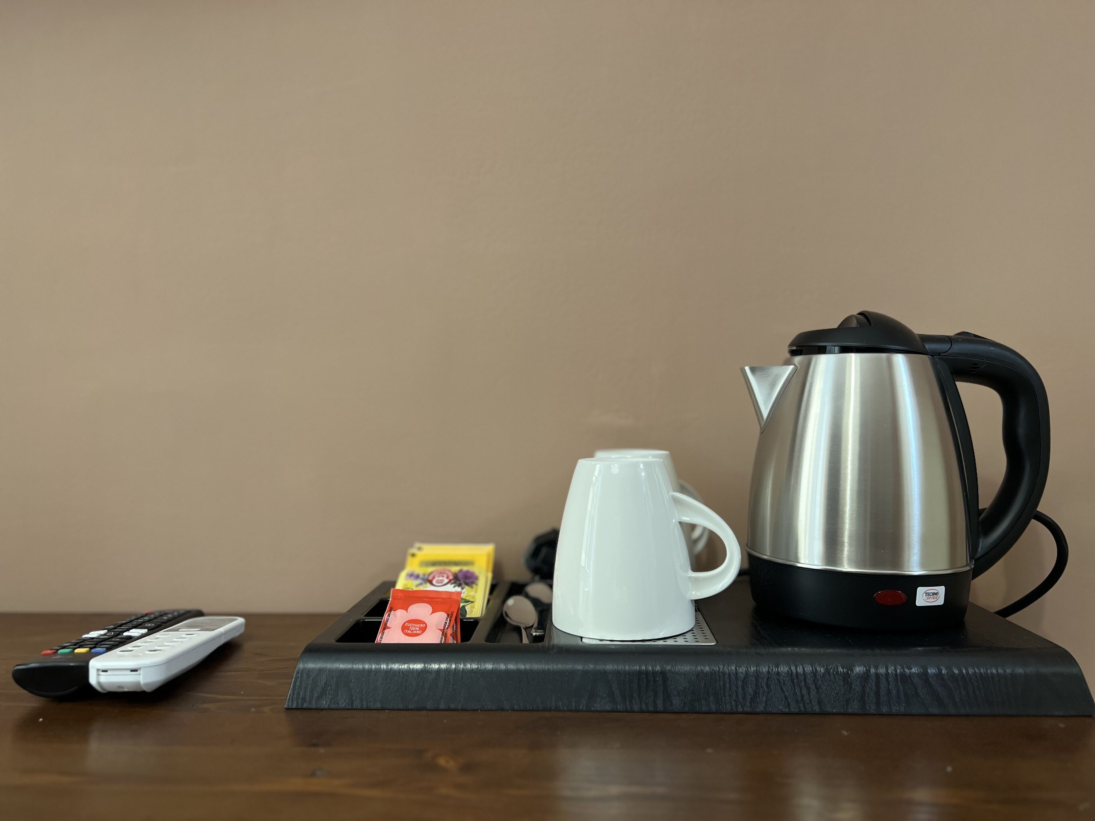 A kitchen counter with a kettle, a white mug, a remote control, and a box of tea.