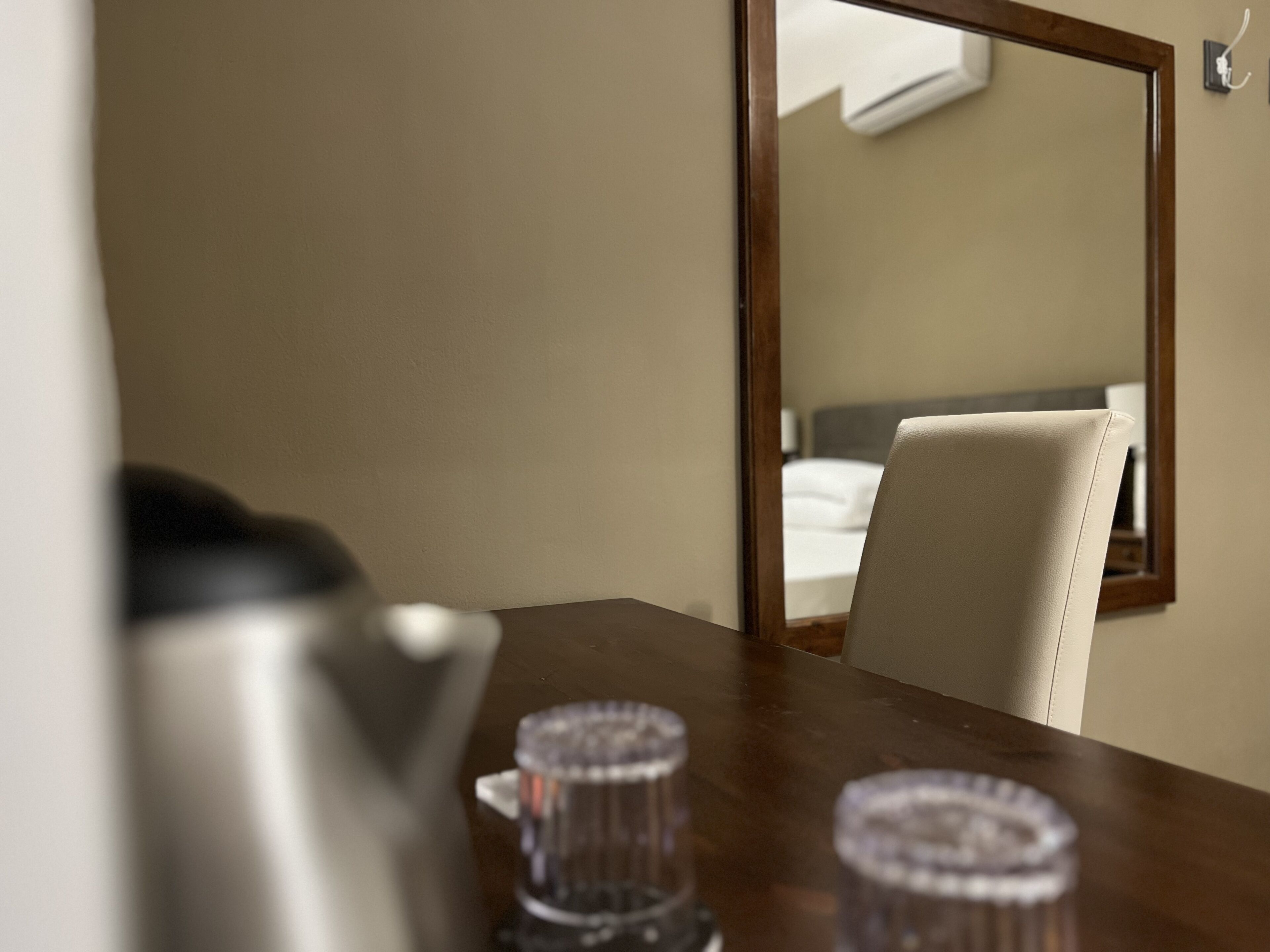 Ein Hotelzimmer mit einem Holztisch, zwei Gläsern, einem Wasserkocher und einem großen Spiegel, der ein Bett und ein Fenster reflektiert.