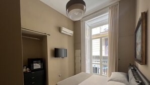 A hotel room with a bed, a TV, an air conditioner, a wardrobe, and a window with a view of buildings. - Relais Ranieri (Naples)
